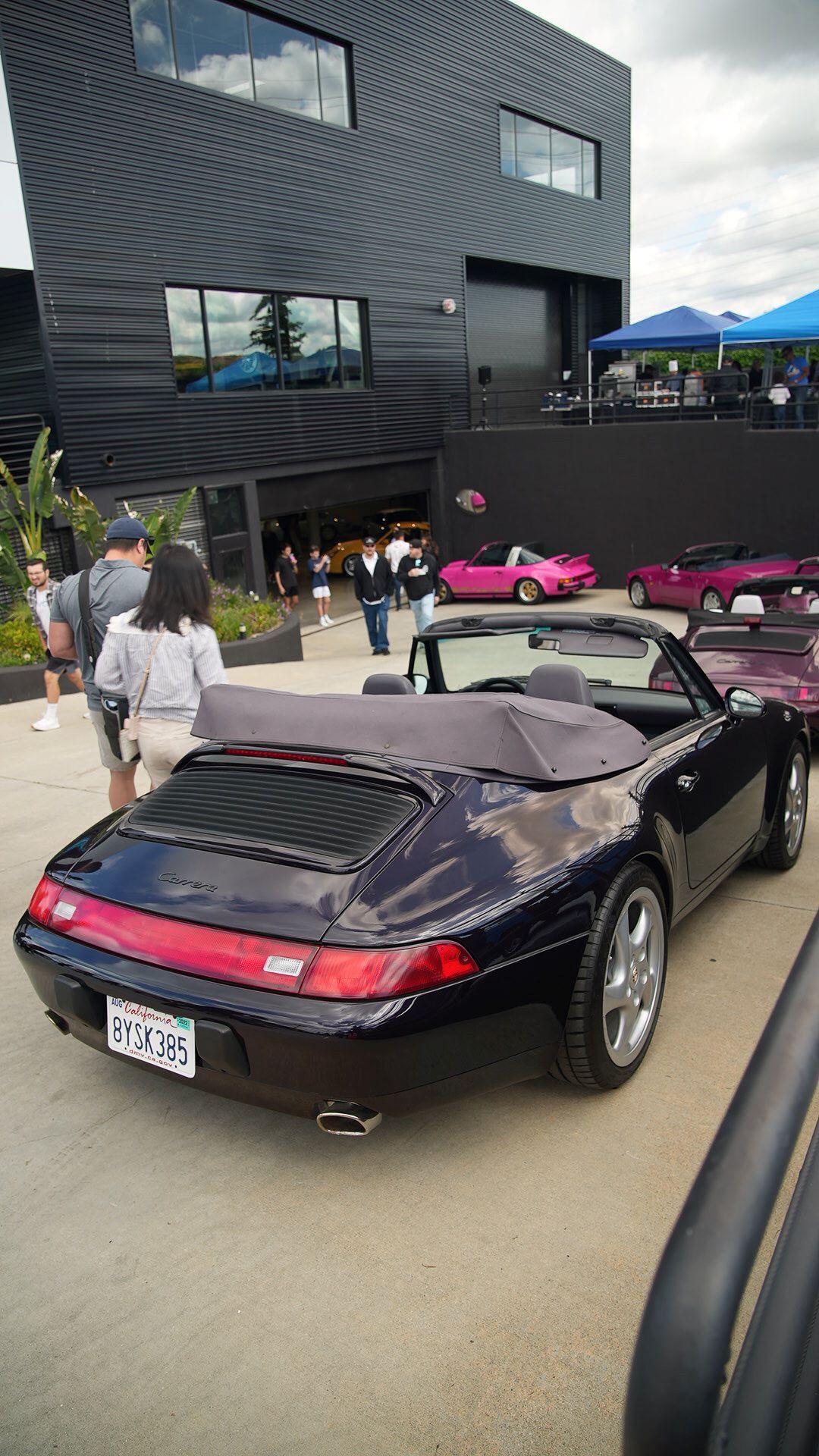 Dark blue Porsche 911 convertible parked outside a black building, with people and other vintage Porsches.