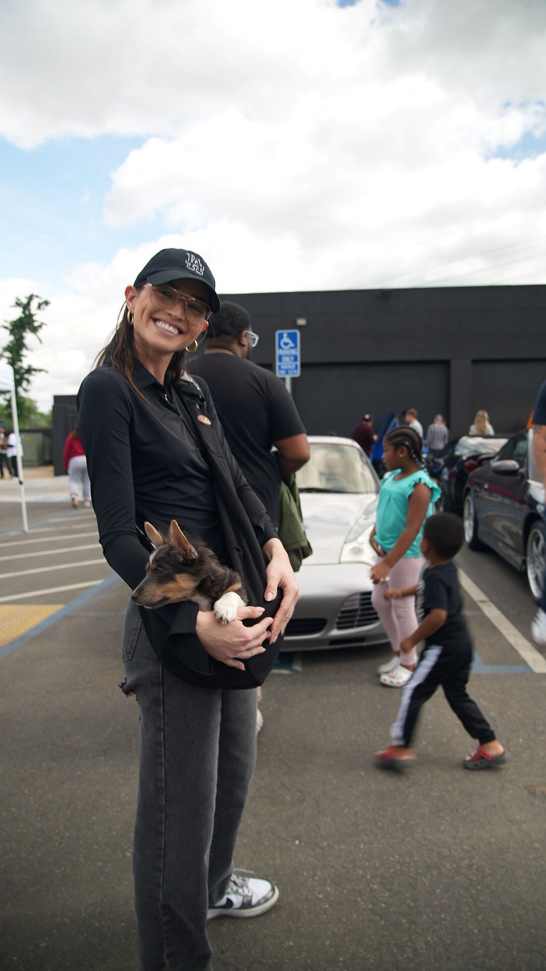 A smiling woman in a black LA Dodgers cap and sunglasses holds a small dog in a black sling.