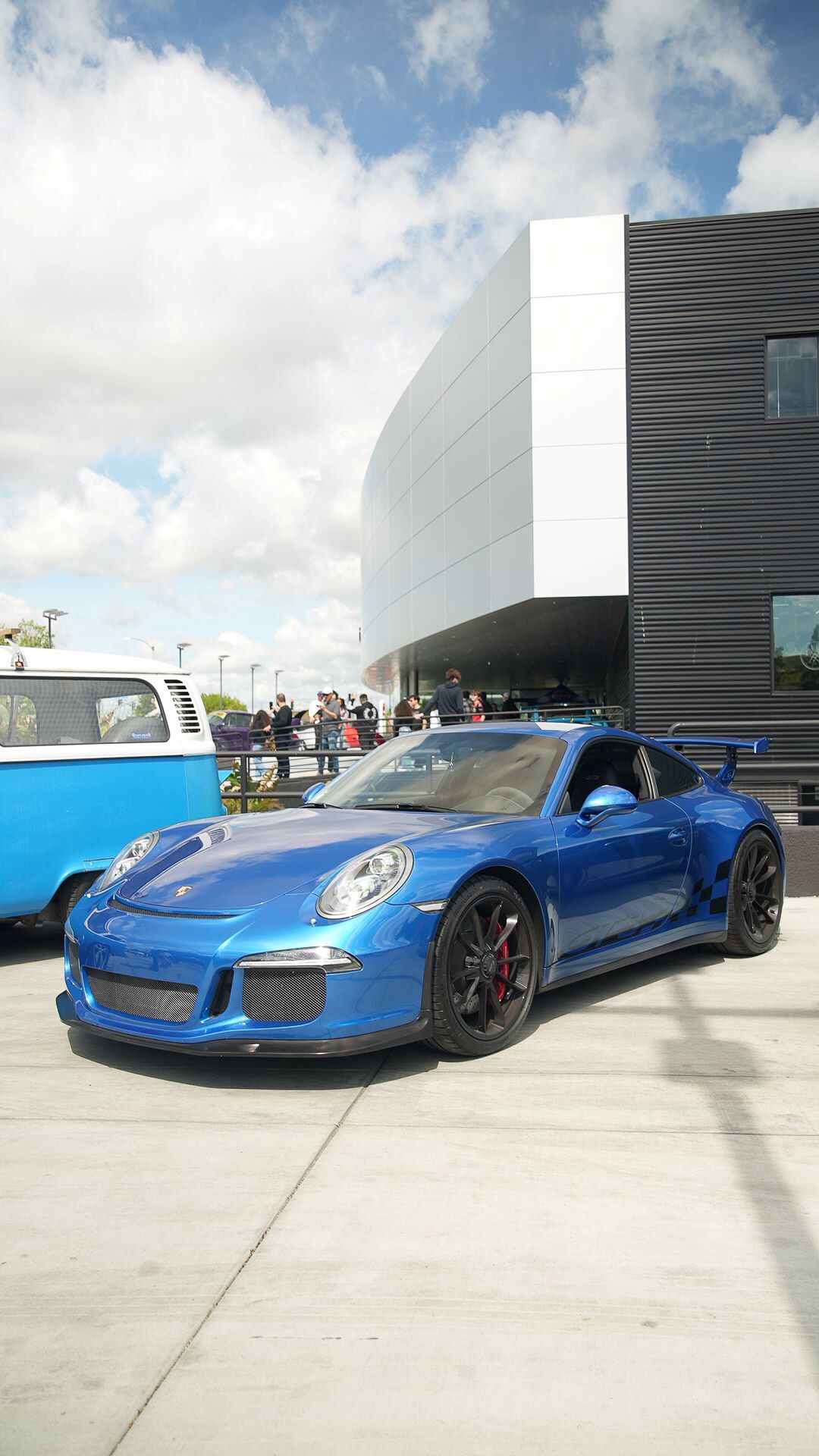 A metallic blue Porsche 911 GT3 with black wheels and red calipers, parked on concrete in front of a modern building.