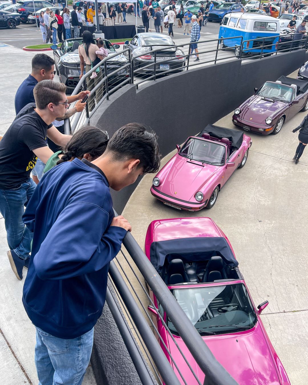 People look down from a ramp at a car show, admiring two pink and a purple vintage convertible Porsches.