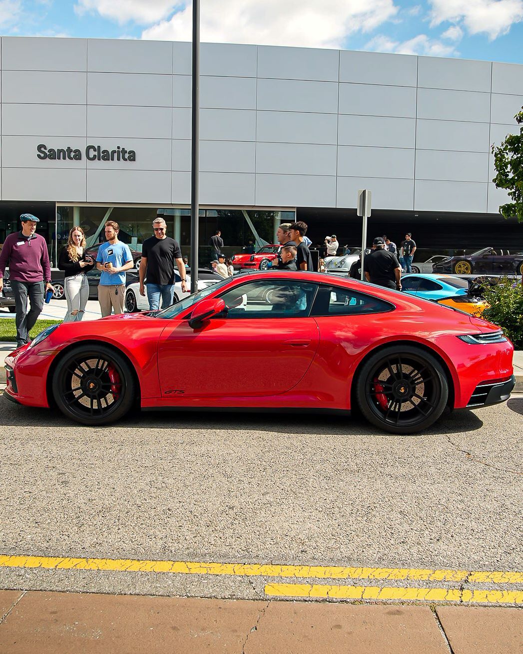 Red Porsche GTS parked on a street with yellow lines, in front of a building labeled 