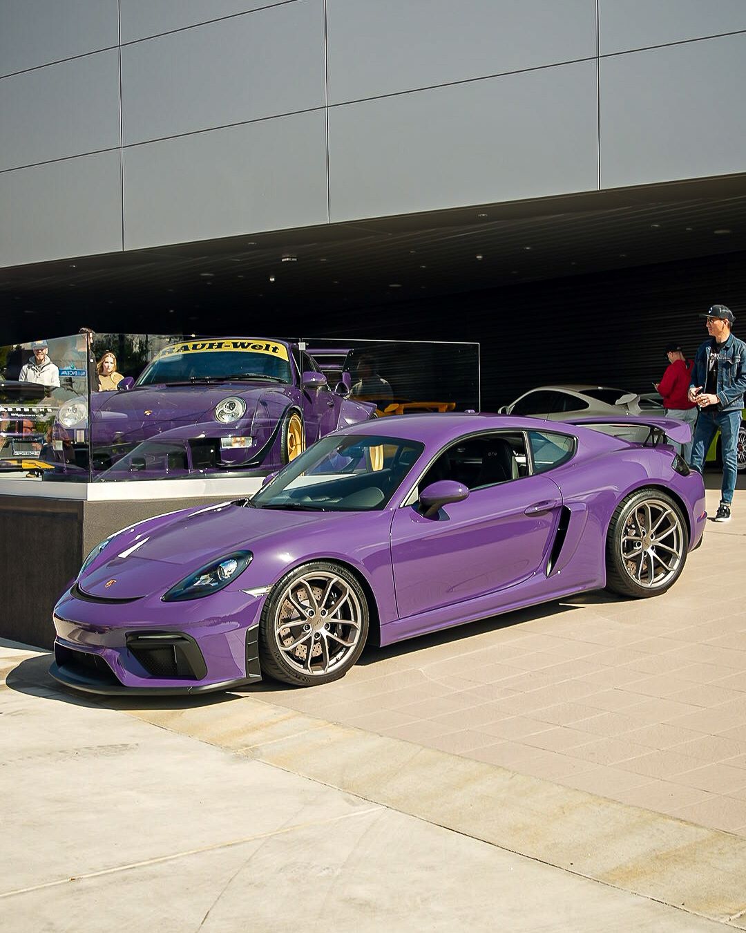 A vibrant purple sports car with silver wheels in the foreground, and a purple Porsche in a glass case behind it.
