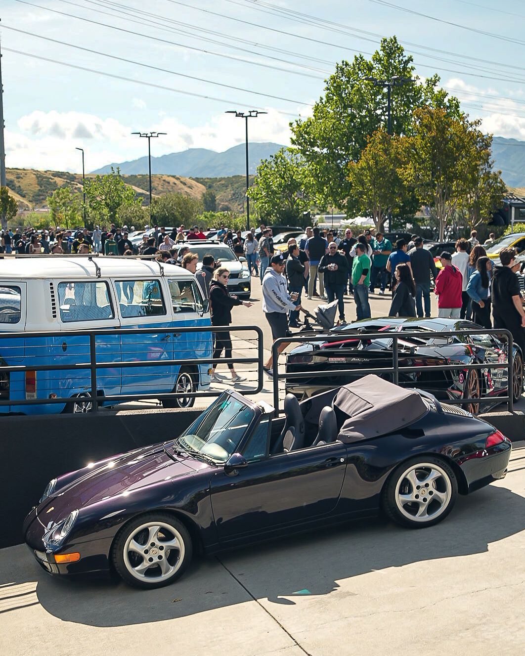A dark purple convertible Porsche in the foreground of a crowded outdoor car show with a blue and white VW bus behind a railing.
