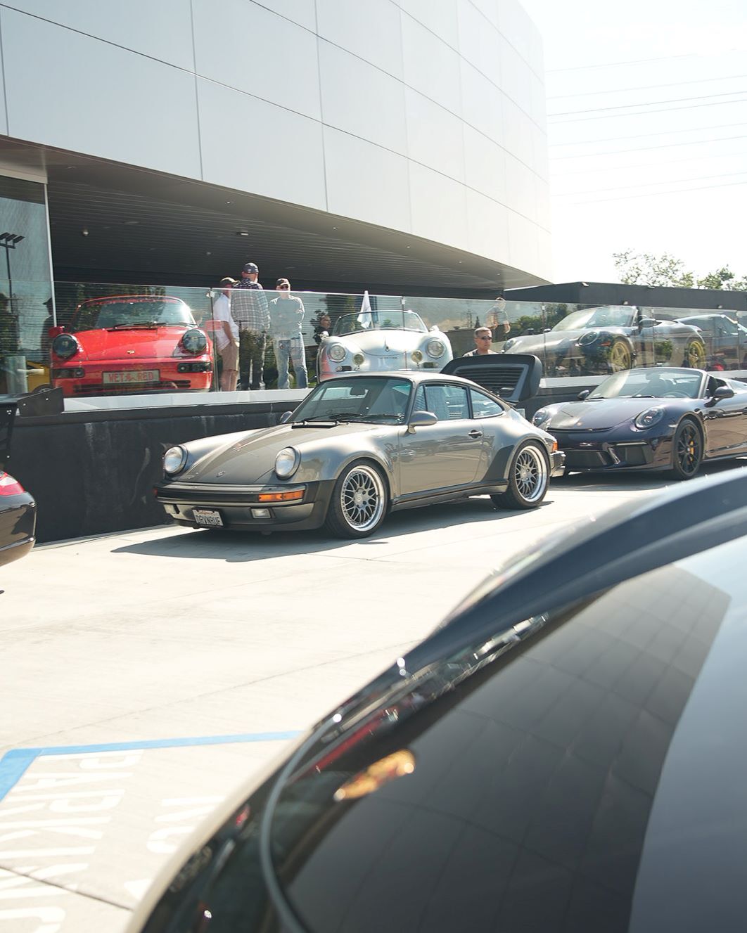 A gray Porsche 911 Targa with its engine cover open, parked on asphalt with other Porsches.