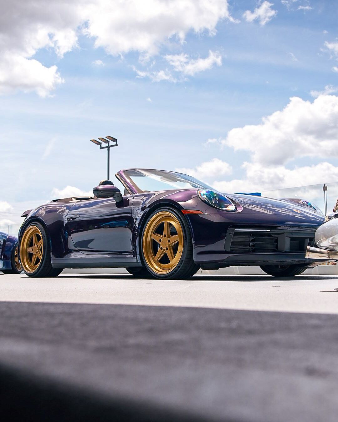 Low-angle view of a shiny purple Porsche convertible with gold rims under a partly cloudy sky.
