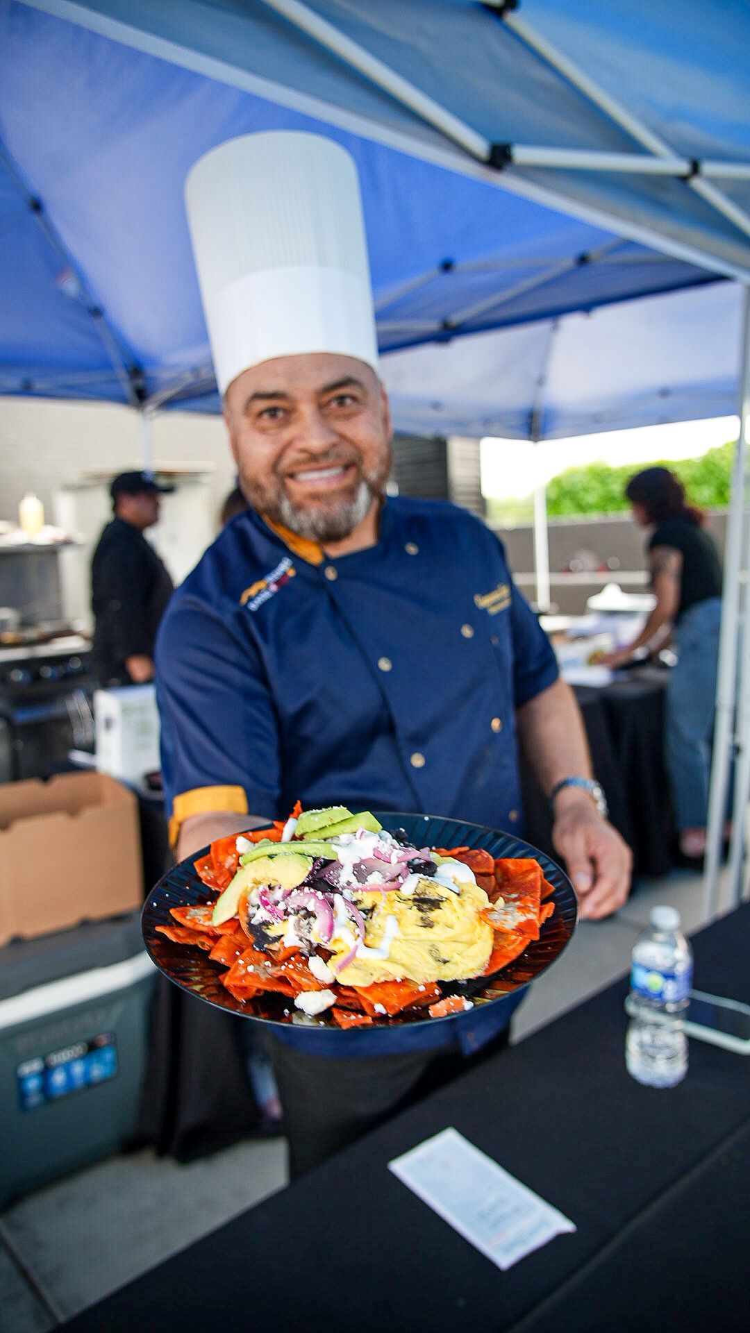 A smiling chef in a tall white hat and blue uniform holds out a plate of chilaquiles topped with avocado and sour cream.