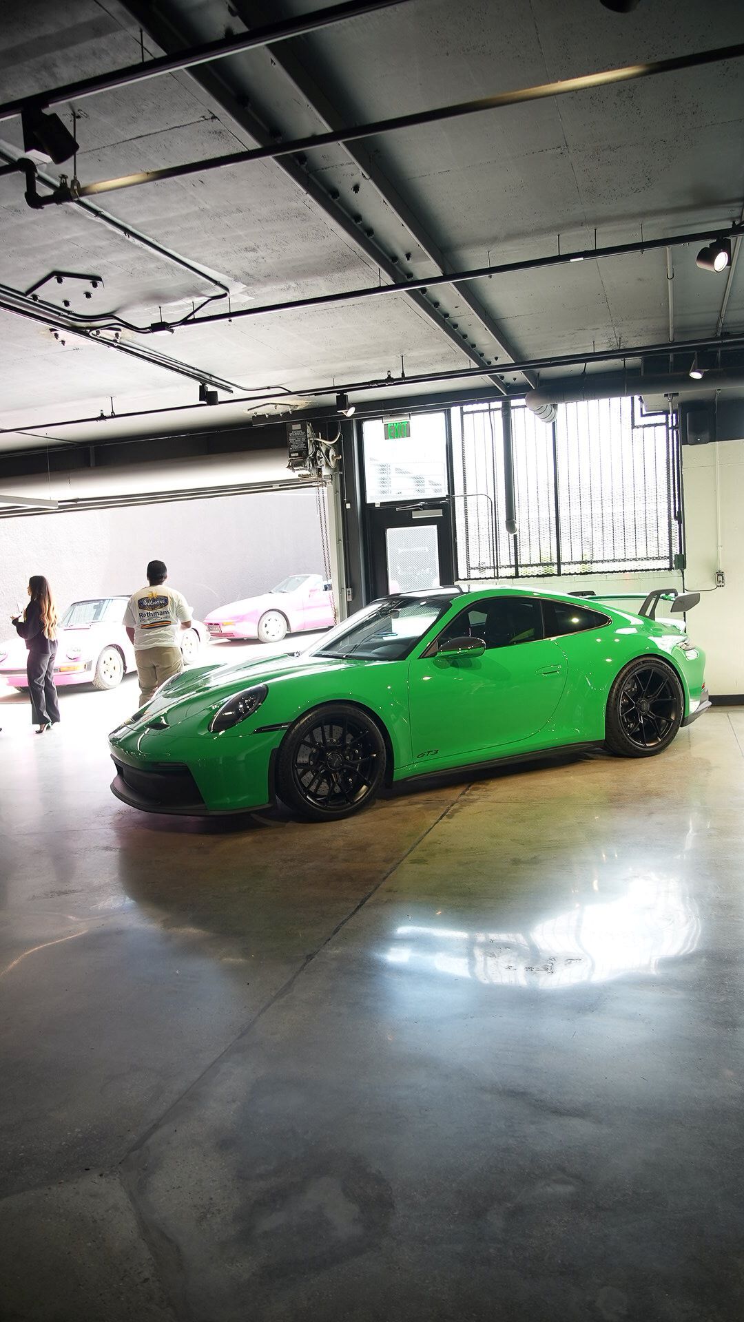 A bright green Porsche sportscar with black wheels parked indoors. Two people stand near two pink Porsches in the background.