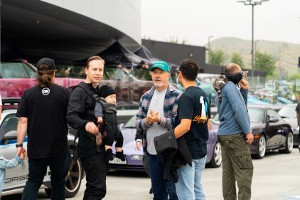 A man holding a baby in a carrier, and three other men, stand and chat around parked sports cars.