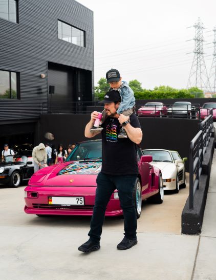 A man carries a child on his shoulders in front of a pink sports car at a car show.