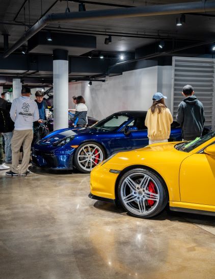 A blue Porsche and yellow Porsche are displayed in a showroom as people observe the vehicles.