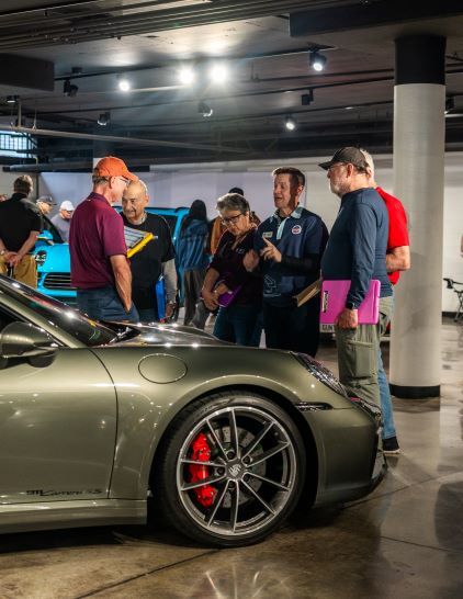 Several people examine a sleek, gray Porsche sports car at an indoor event with bright overhead lighting.