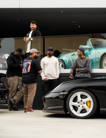 Four people converse in front of a sleek, turquoise Porsche and a black car in a showroom.