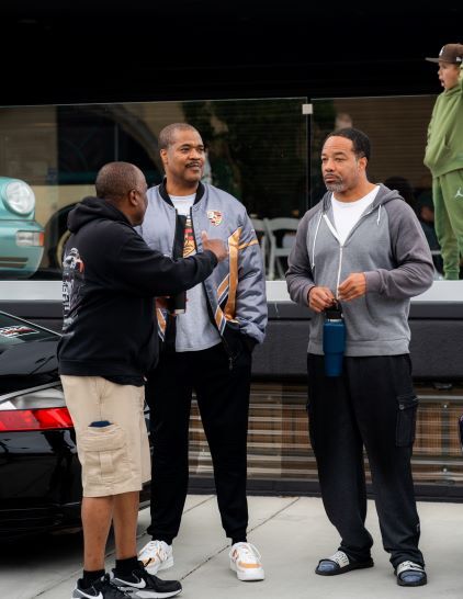 Three men in casual clothing converse and smile while standing near a vintage car.
