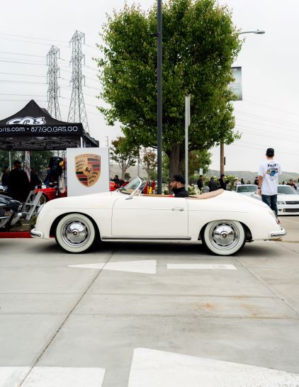 A classic white Porsche convertible sits parked at an outdoor car show event.