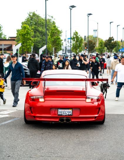 A bright red Porsche with a large spoiler sits on a city street surrounded by a crowd of people and a dog.