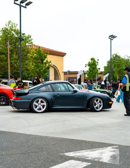 A dark green Porsche 911 sits parked at a car show with people admiring it.