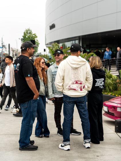 Several people converse outdoors near a modern building and a classic red car.