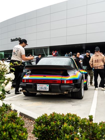 A classic Porsche with a rainbow stripe sits parked as people gather around it at an event.