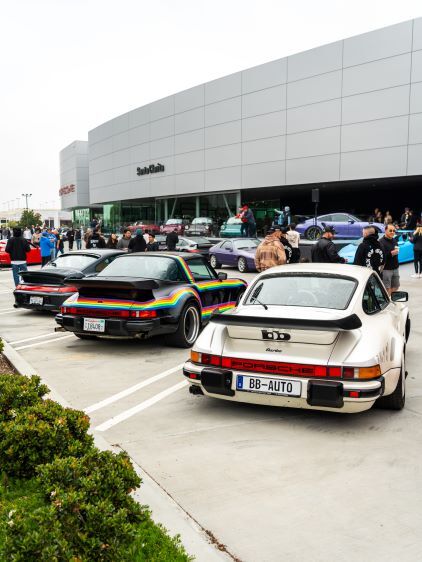 Classic Porsche cars, including a striped model, are parked in front of a modern Porsche dealership.