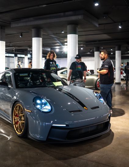 Three people examine a gray Porsche sports car displayed in a well-lit garage.
