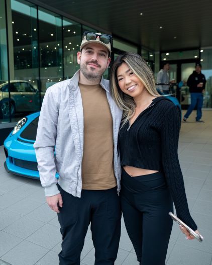 A man and woman smile while posing for a photo in front of a blue sports car and modern building.