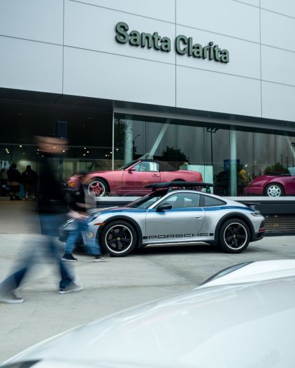 A silver Porsche convertible sits outside a Santa Clarita dealership as blurred pedestrians walk past.