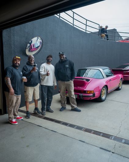 Four men pose with a vintage pink Porsche in a garage setting.