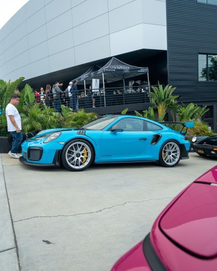 A bright blue Porsche 911 GT2 RS sits parked at an outdoor car show with people observing.