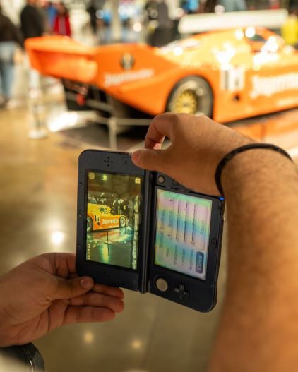 Hands hold a Nintendo DS displaying a picture of an orange race car, with the actual car blurred in the background.