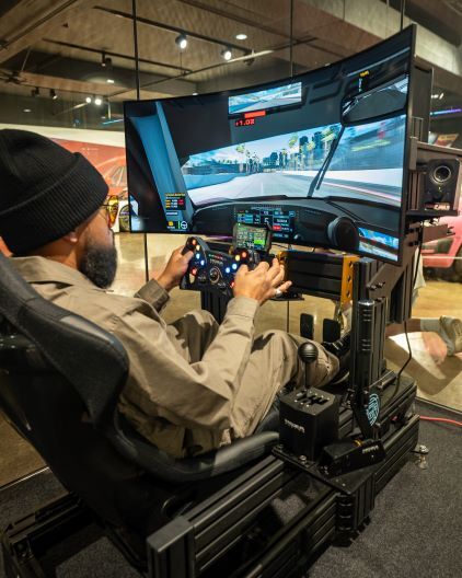 A man wearing a beanie and sunglasses plays a racing game on a simulator with a steering wheel and pedals.