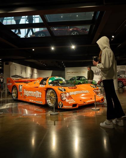 A person takes a photo of an orange Porsche race car displayed in a museum.