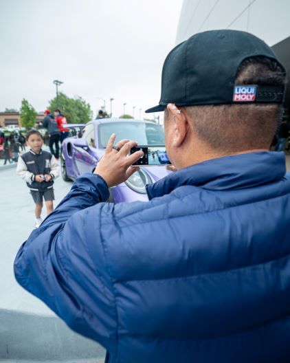 A person in a blue jacket and black hat taking a photo of a child and a purple sports car with a smartphone.