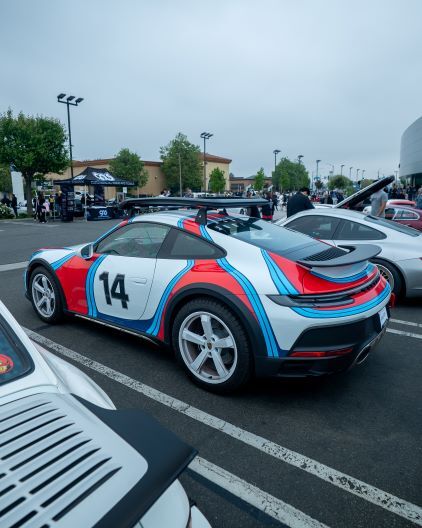 A white, red, and blue Porsche 911 GT3 with racing stripes sits parked in a crowded lot.