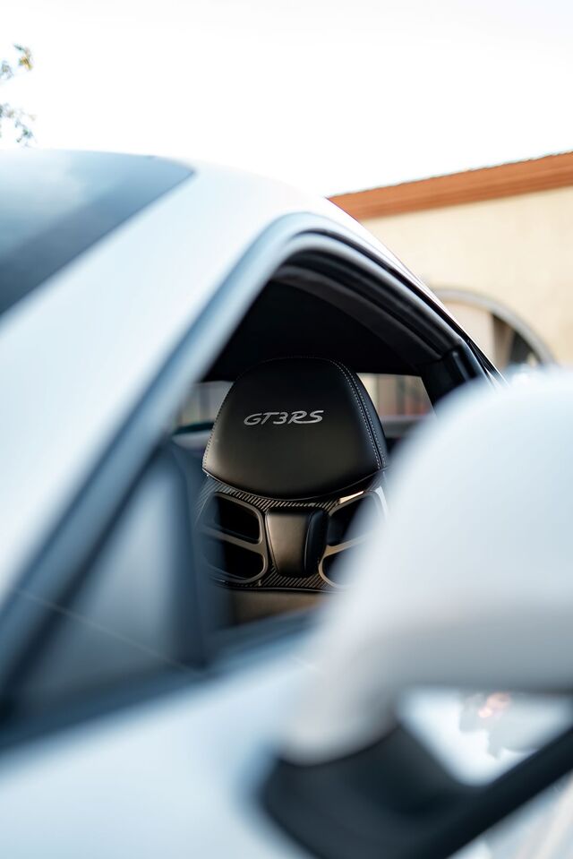 A stark white Porsche GT3 RS with black interior seats visible through the window, with 'GT3RS' embroidered on the headrest.