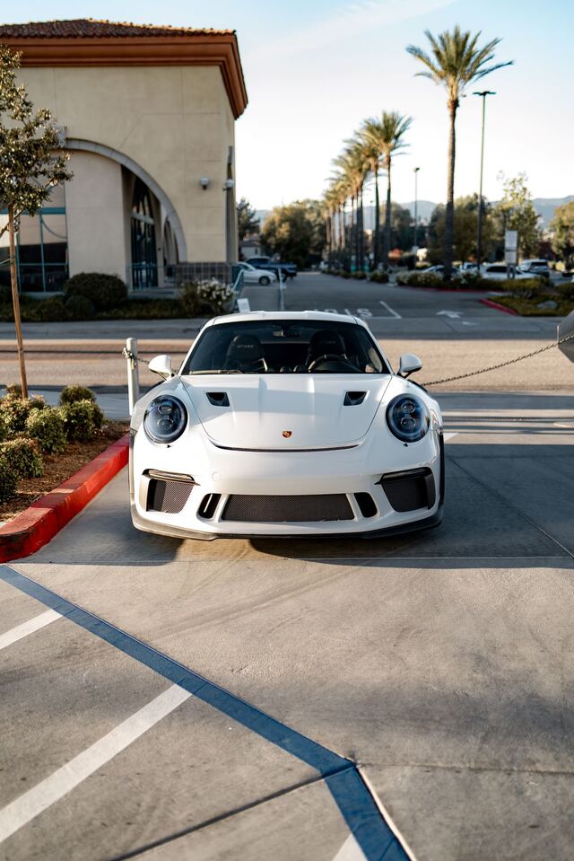 Front view of a white Porsche 911 parked on concrete, with palm trees and a building in the background.