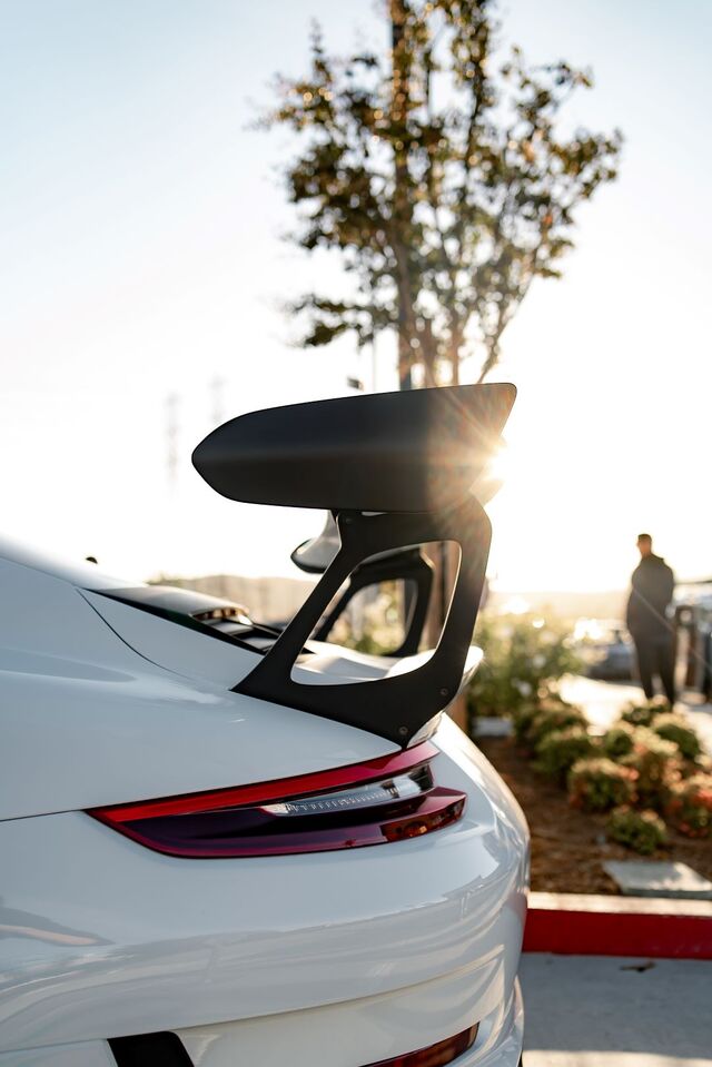 Rear quarter view of a white sports car with a prominent black spoiler, sunlight flaring over it.
