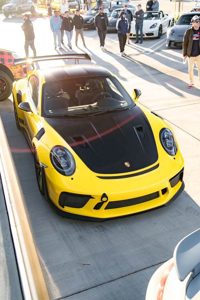 A bright yellow Porsche 911 with a black hood sits at a car show, surrounded by onlookers.