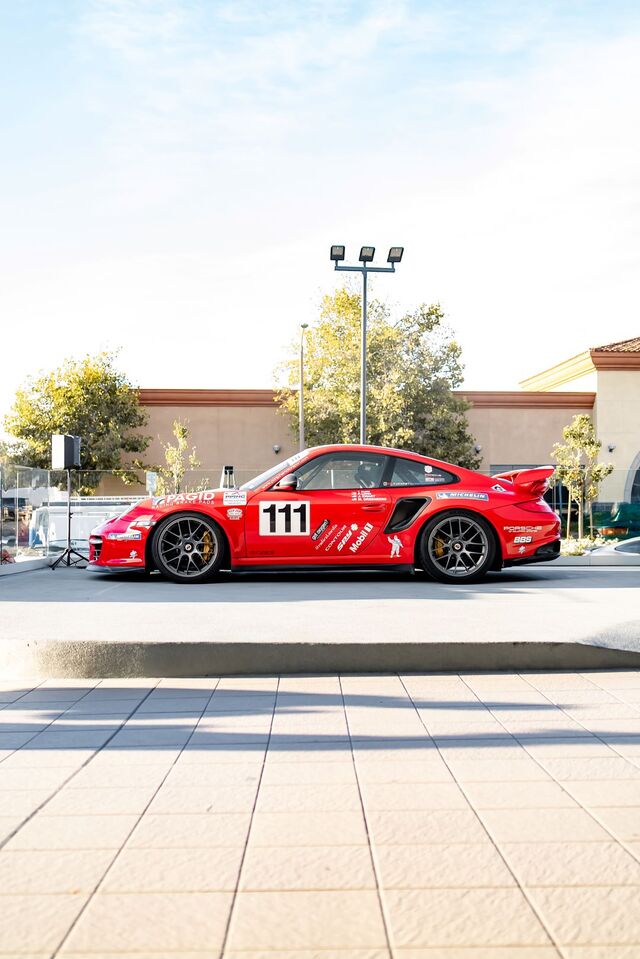 Red Porsche race car #111 with sponsor decals, parked on a light-colored paved lot.