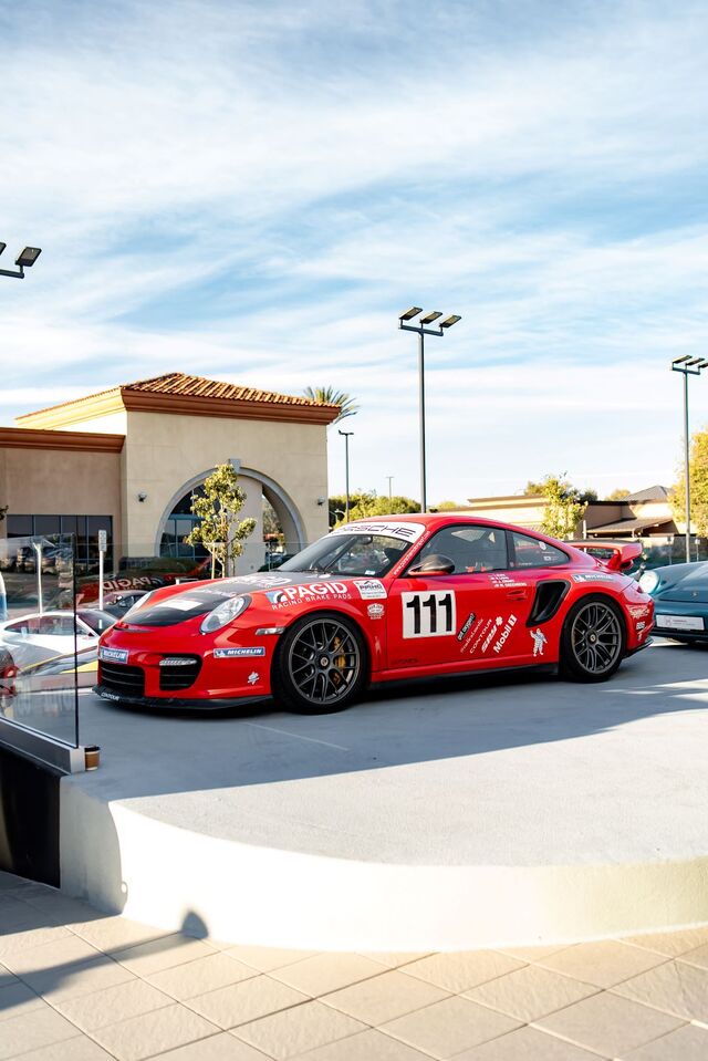 A red Porsche race car with number 111 sits in a parking lot under a partly cloudy sky.