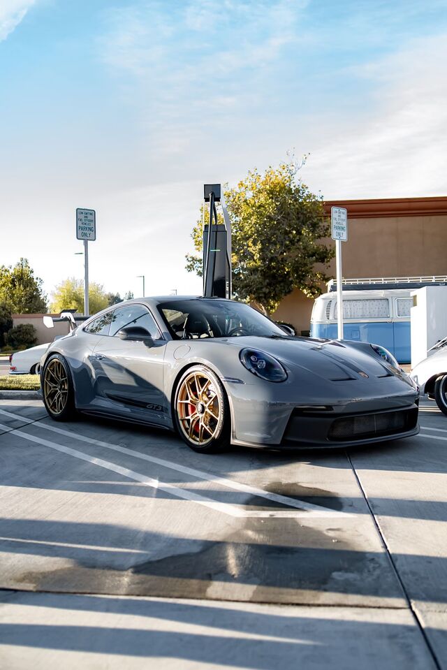 A gray Porsche 911 GT3 with gold wheels parked in a lot beside a charging station and an old VW van.