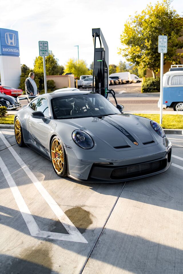 A gray Porsche 911 GT3 with gold wheels parked in a lot beside a charging station and an old VW van.