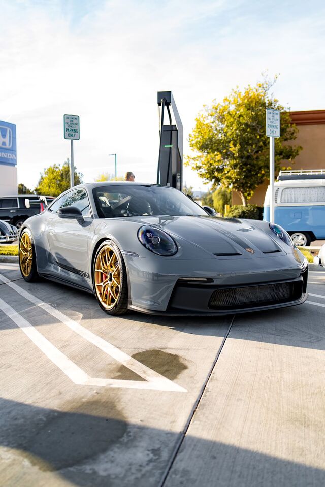 A gray Porsche 911 GT3 with gold wheels sits parked near an electric vehicle charging station.