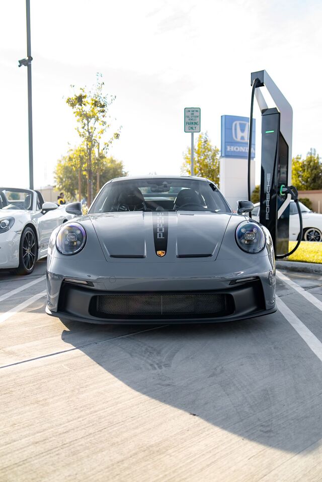 A gray Porsche sports car with a black racing stripe down the hood is parked next to an EV charging station.