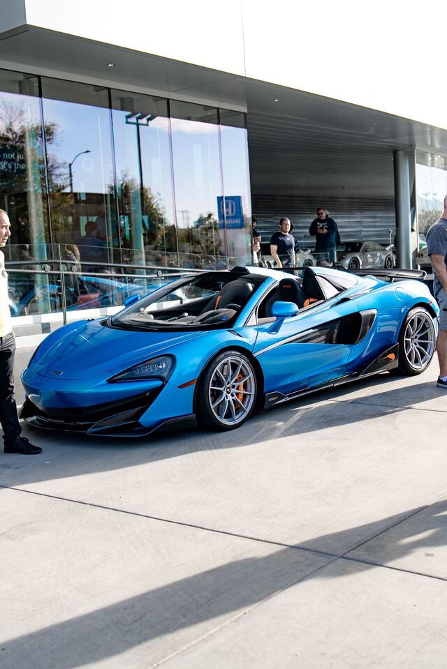 A highly descriptive image of a convertible blue McLarens parked outside a building with glass doors, seen from a front corner view.