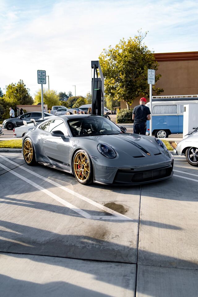 A gray Porsche 911 GT3 with gold wheels parked in a lot beside a charging station and an old VW van.
