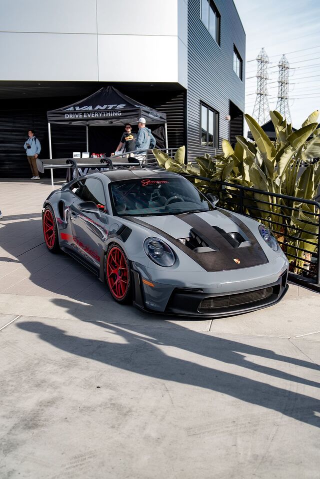A grey Porsche 911 GT3 RS with red wheels and decals on a polished concrete floor in a showroom with people.