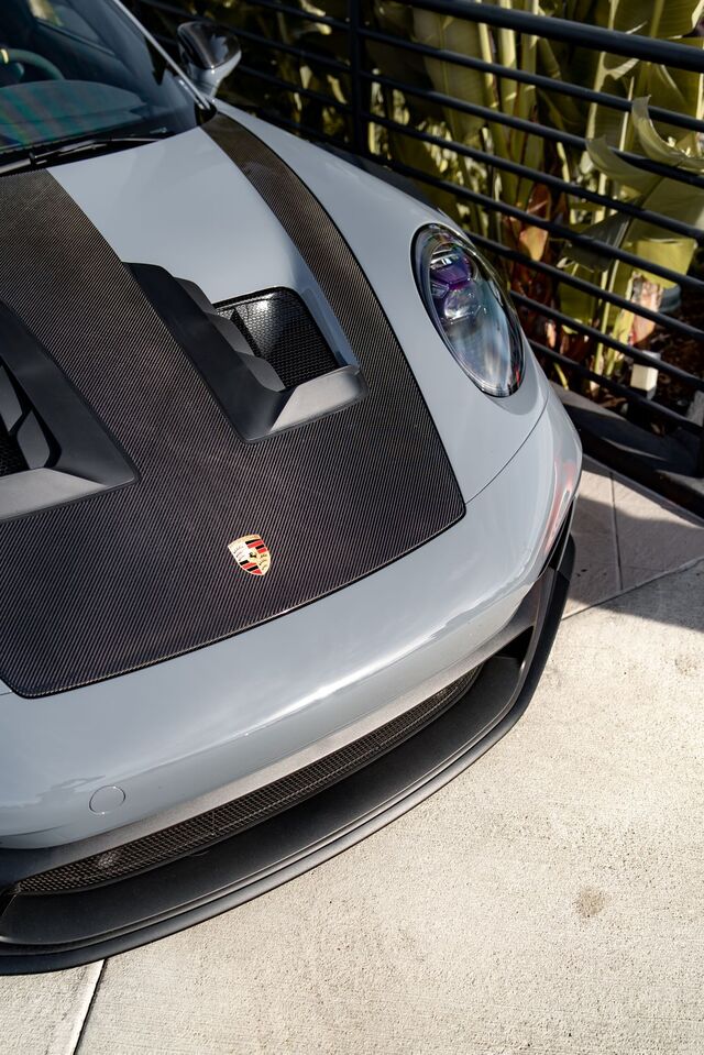 A silver Porsche with a carbon fiber hood and logo sits parked on a concrete surface.