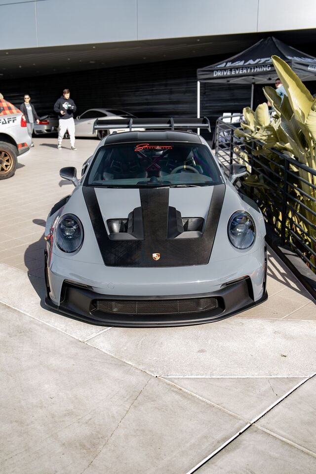 A front view of a gray Porsche 911 parked outdoors, featuring a black racing stripe on its hood.