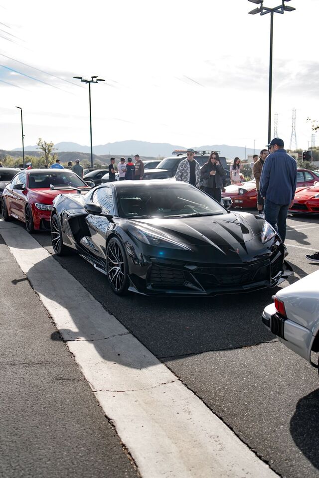 A sleek black Corvette and a red sports car parked in a lot, with people and mountains in the background.