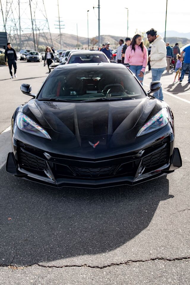 Front view of a sleek black Corvette Z06 with colorful headlights, parked, with people in the background.